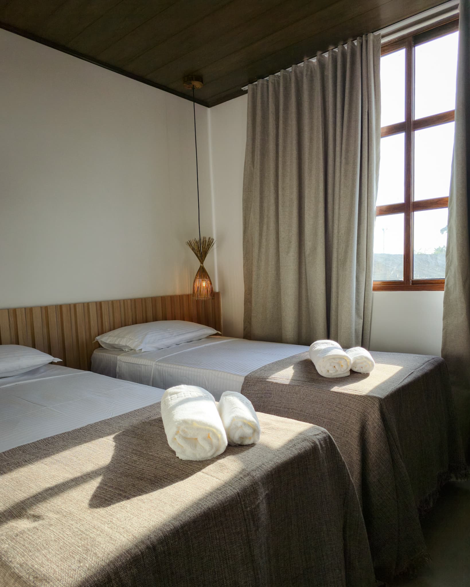 Two twin beds with rolled white towels in a sunlit bedroom with beige curtains.