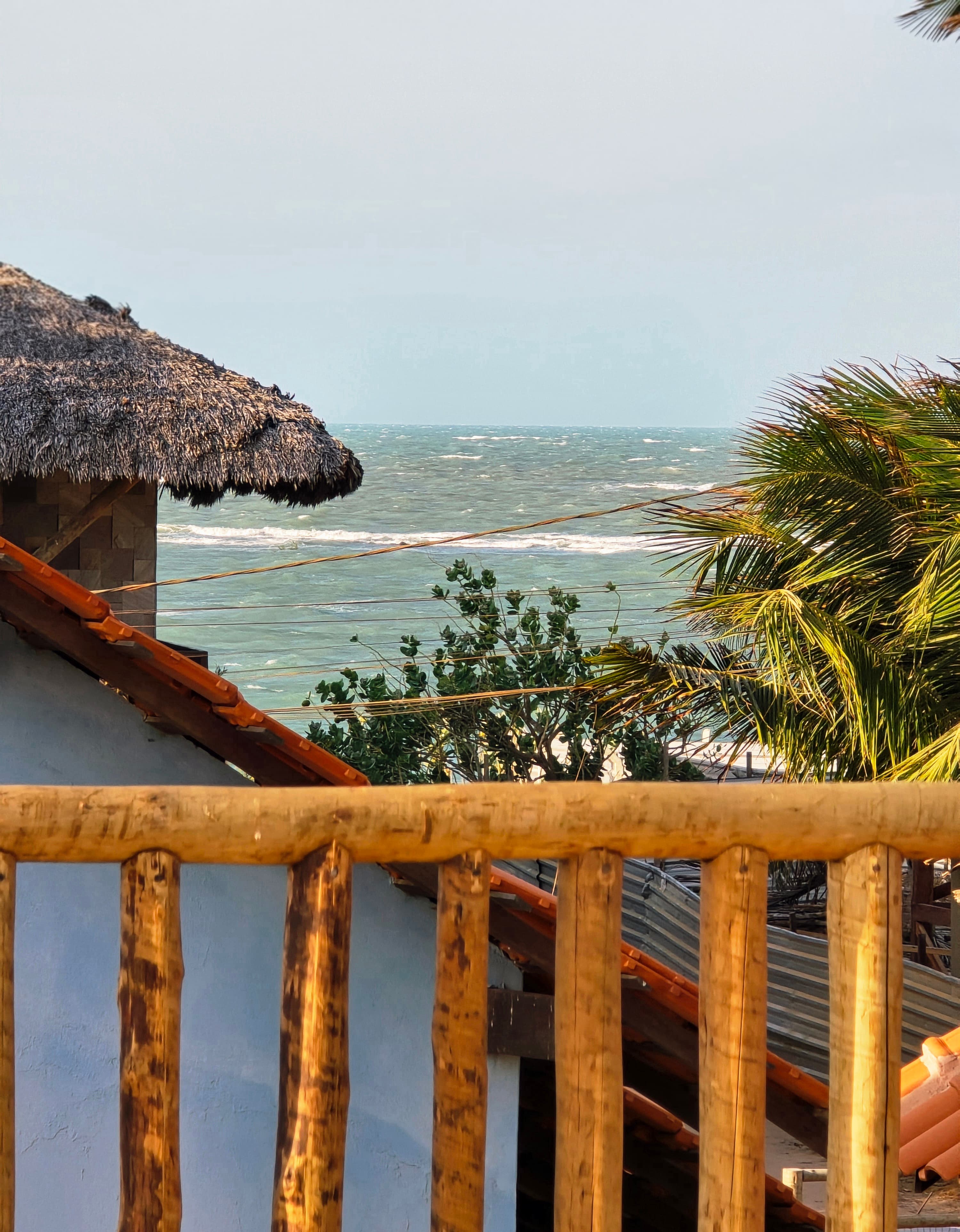 Wooden balcony railing overlooking a tropical ocean scene with thatched roofs and palm trees.