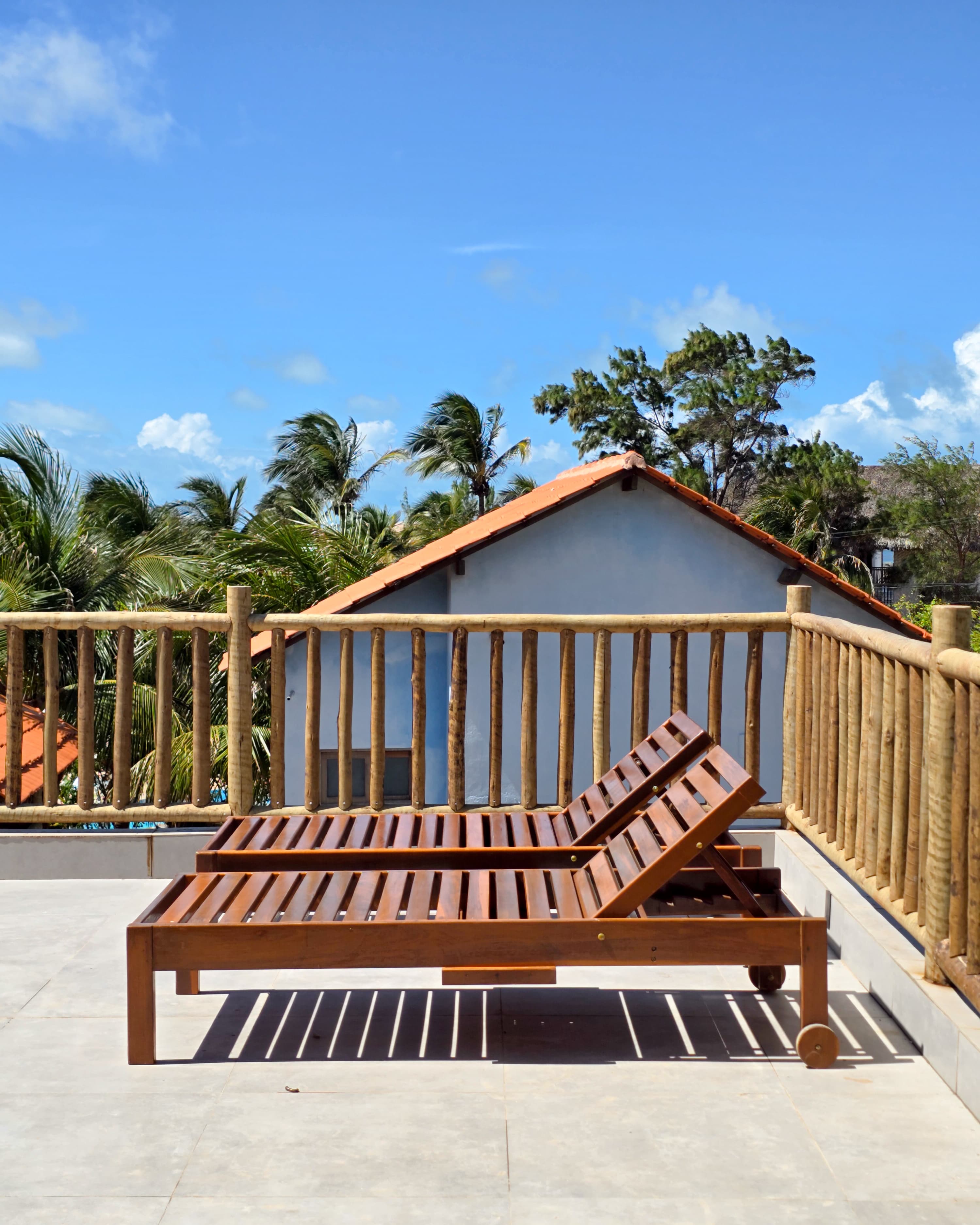 Two wooden lounge chairs on a sunny balcony overlooking palm trees and blue sky.