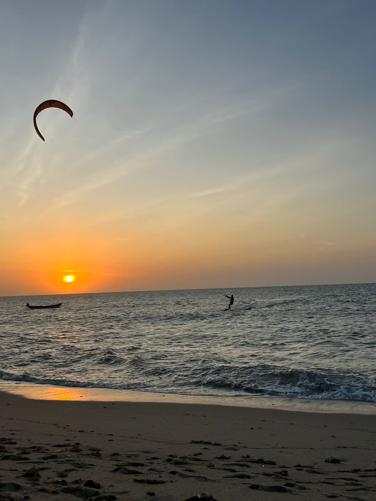Kitesurfer silhouetted against a vibrant orange sunset over the ocean with a small boat.